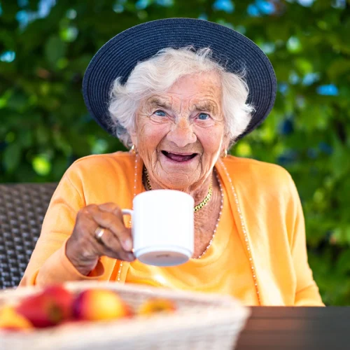 Klientin draußen am Tisch mit Becher in der Hand sitzend und in die Kamera lachend. Im Hintergrund Blätter verschwommen, im Vordergrund auf dem Tisch ein Korb mit Äpfeln verschwommen.