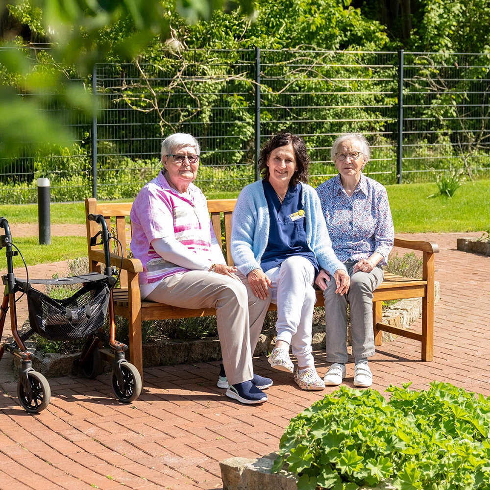 1 Pflegekraft in der Mitte und zwei Klientinnen draußen auf einer Bank auf einem gepflasterten kleinen Platz sitzend, drumherum Büsche und Bäume an einem Zaun
