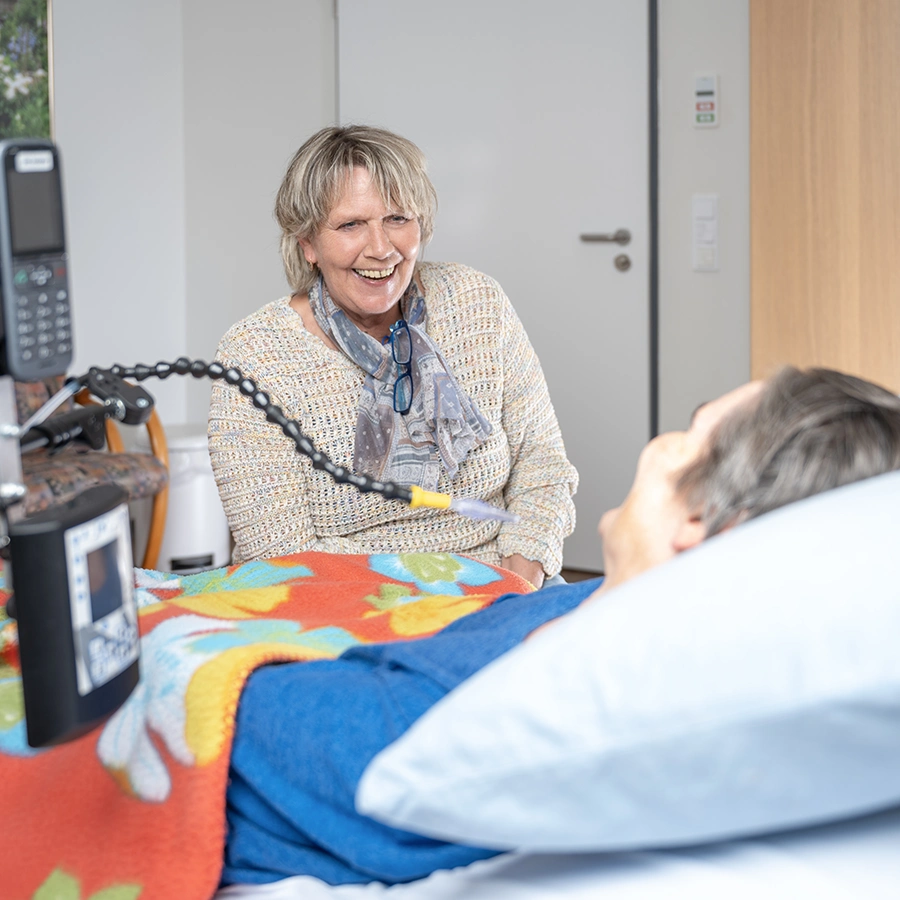 Eine weibliche Person sitz am Bett von Frau Salzmann und lacht sie an. Im Vordergrund leicht verschwommen und Aufnahme von hinten im Bett liegend, den Kopf zur Person gedreht und links am Rand vom Bild eine Sprachsteuerung auf einem Schwenkarm.