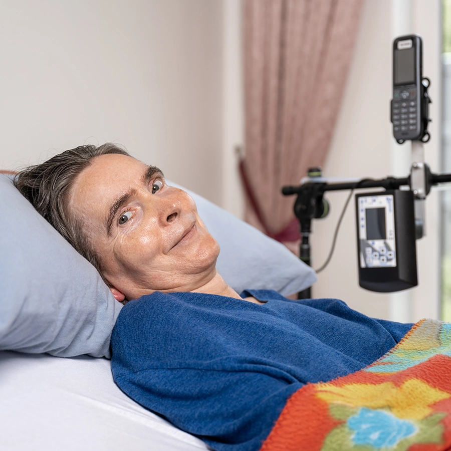 Frau salzmann liegt im Bett, lächelt in die Kamera und eine Sprachsteuerung auf einem Schwenkarm ist über ihrem Bett