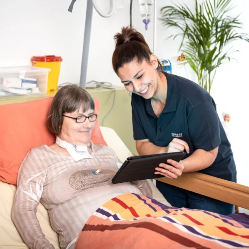 Pflegekraft am Bett einer Klientin zeigt auf ein Tablet, auf das bei schauen.