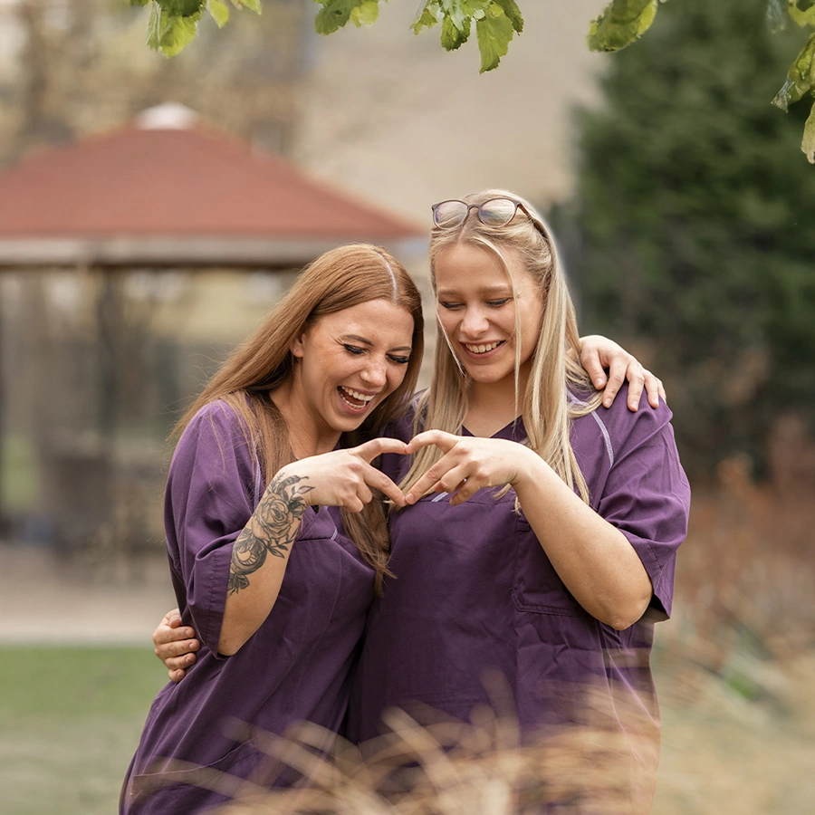 2 Pflegerinnen draußen auf einer Wiese umarmen sich und zeigen ein Herz mit ihren Händen zusammen. Beide schauen darauf und lachen.