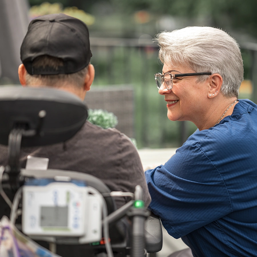 Pflegekraft im Profil hockt am Rollstuhl neben einem Klienten, der von hinten zu sehen ist. Im Hintergrund verschwommen ein Balkon.