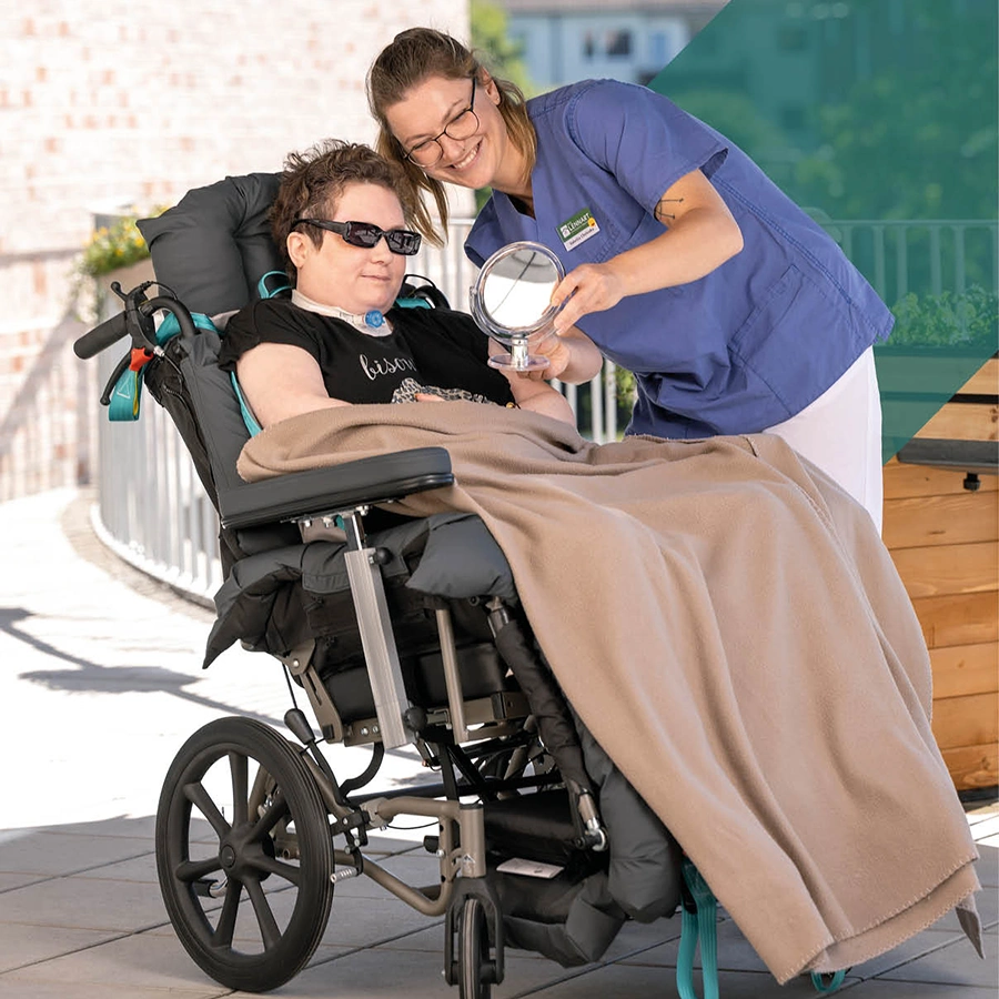 Klientin im Rollstuhl, daneben eine Pflegekraft. Beide schauen in einen Spiegel.