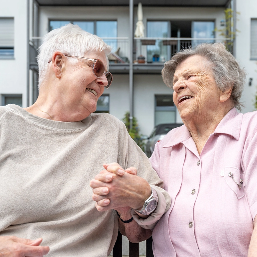 2 Klientinnen sitzen, sich an der Hand haltend, auf einer Holzbank im Garten und lachen sich an.