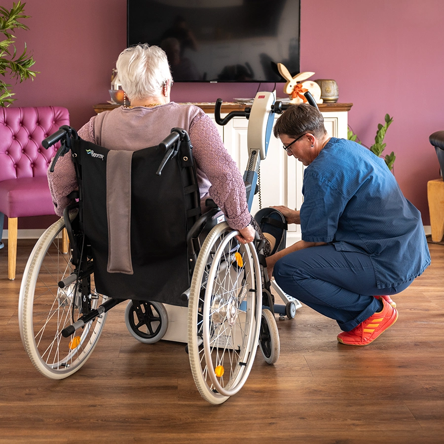 Klientin im Rollstuhl mit Ansicht von hinten. Daneben kniet eine weibliche Pflegekraft in Seitenansicht, welche am Rollstuhl in Fußhöhe eine Einstellung vornimmt.