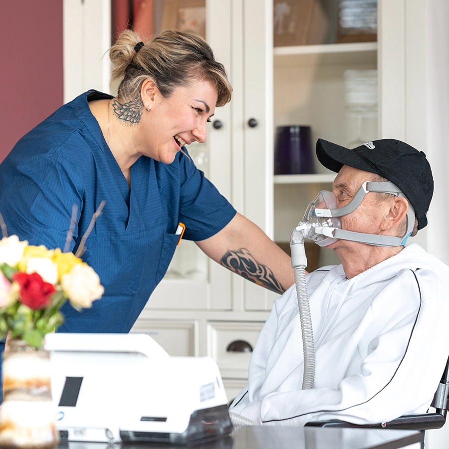 Klient auf einem Rollstuhl im Zimmer lächelt eine weibliche Pflegekraft an, die neben ihm steht und zurücklächelt.