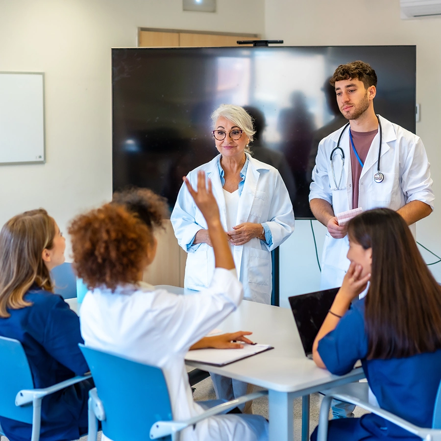 Ärztin und Arzt in Frontansicht steht vor sitzenden Ärztinnen, wobei sich eine gerade meldet.