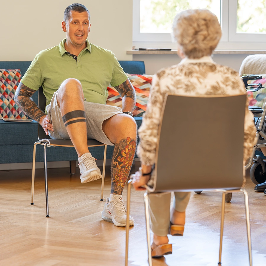 df-leistungen-physiotherapie-02 Trainer macht sitzend eine Physioübung vor. Ein Bein ist angehoben. Davor in Rückansicht eine Klientin sitzend und ihn anschauend.