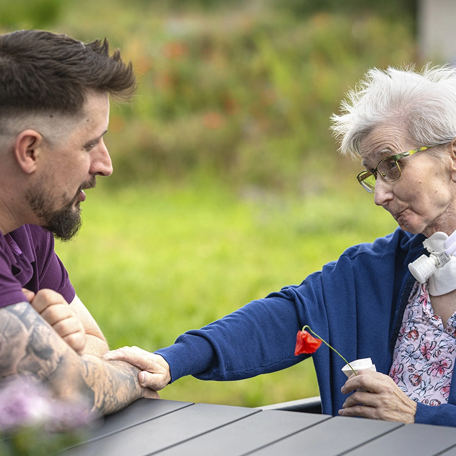 Eine Klientin im Rollstuhl und ein Pfleger sitzen draußen an einem Tisch, im Hintergrund eine Wiese verschwommen dargestellt. Sie hat ein Blume in der Hand, die andere auf des Pflegers Arm gelegt. Beide schauen sich an.