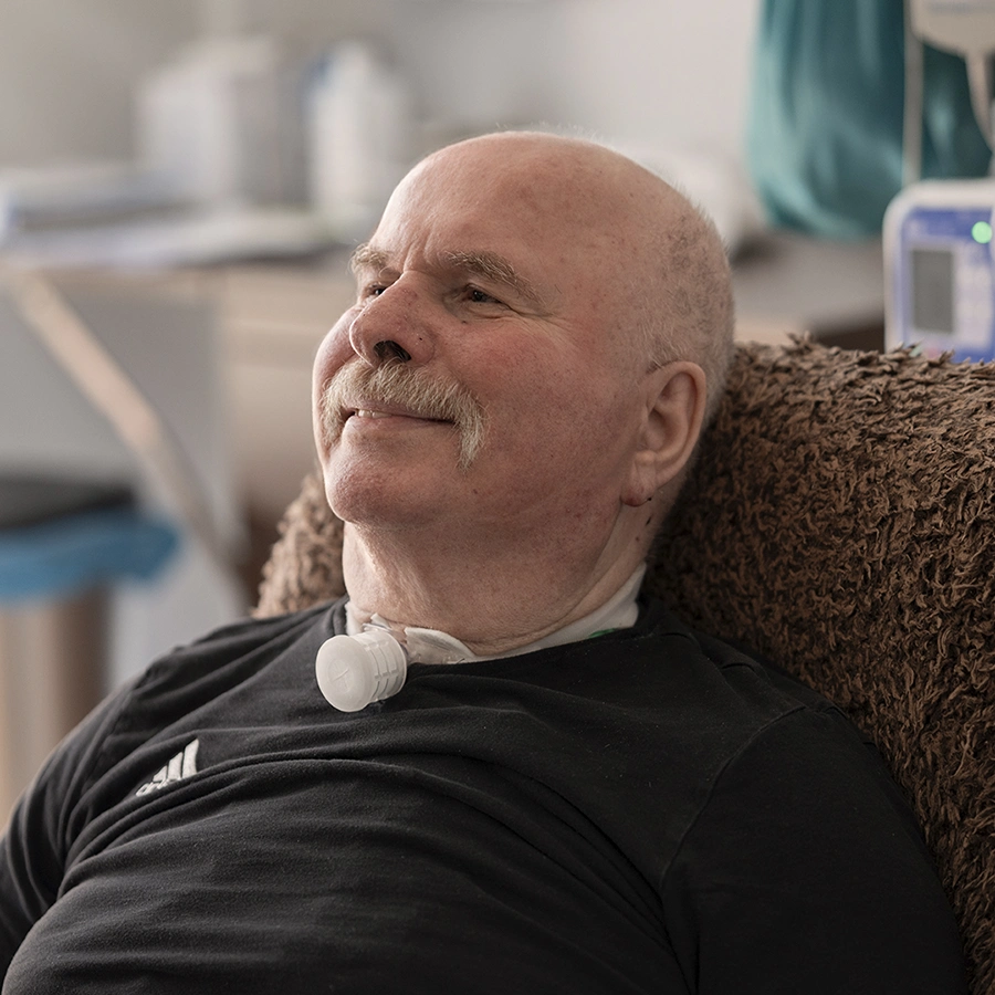 Klient in Schrägansicht auf einem Stuhl sitzend und lächelnd. Im Hintergrund verschwommen eine Ges undheits-Apparatur.