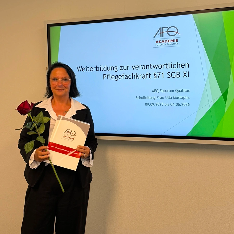 Pflegekraft hält Auszeichnung und eine Rose in den Händen und steht neben einem Wandbildschirm.