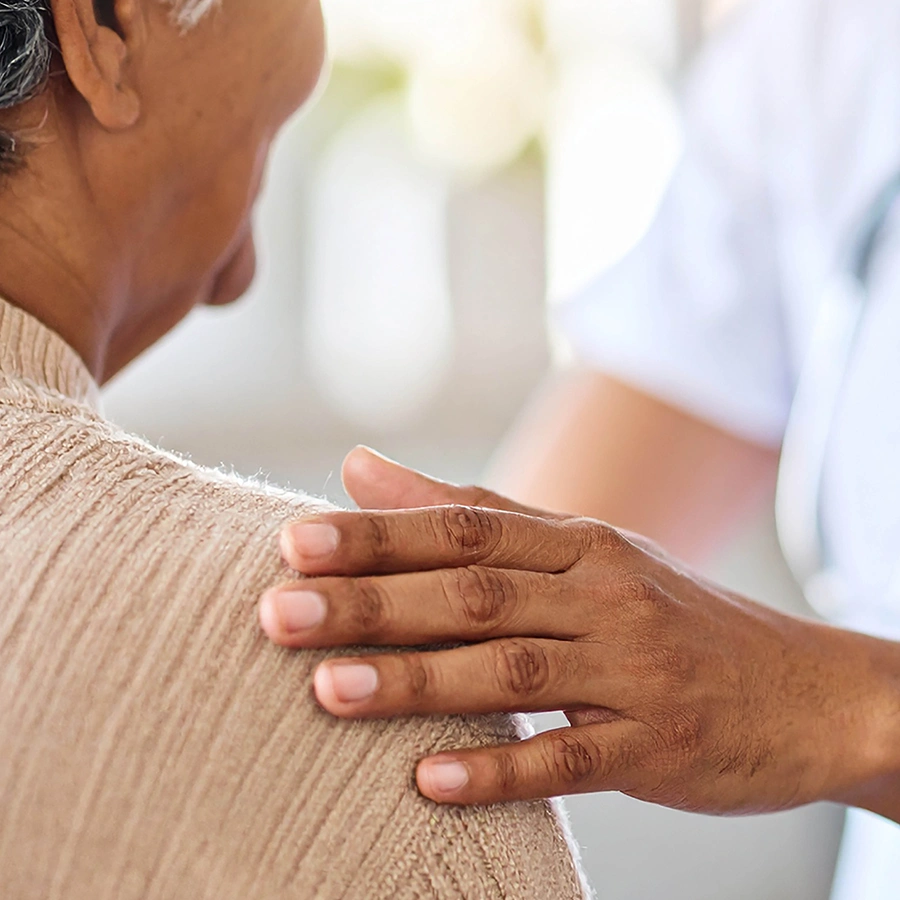Pflegekraft hat eine Hand auf die Schulter einer Klientin gelegt. Im Fokus nur die Hand und der Arm.