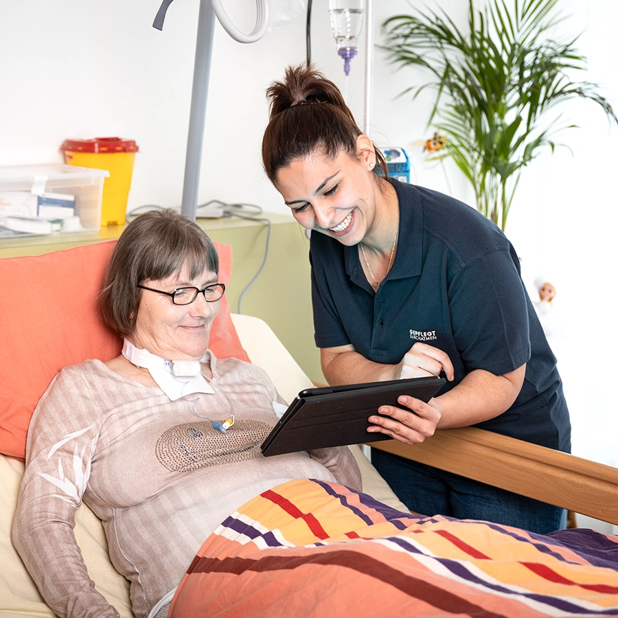 Weibliche Pflegekraft steht am Bett einer Klientin. Beide schauen lächelnd auf ein Tablet in der Hand der Pflegekraft.