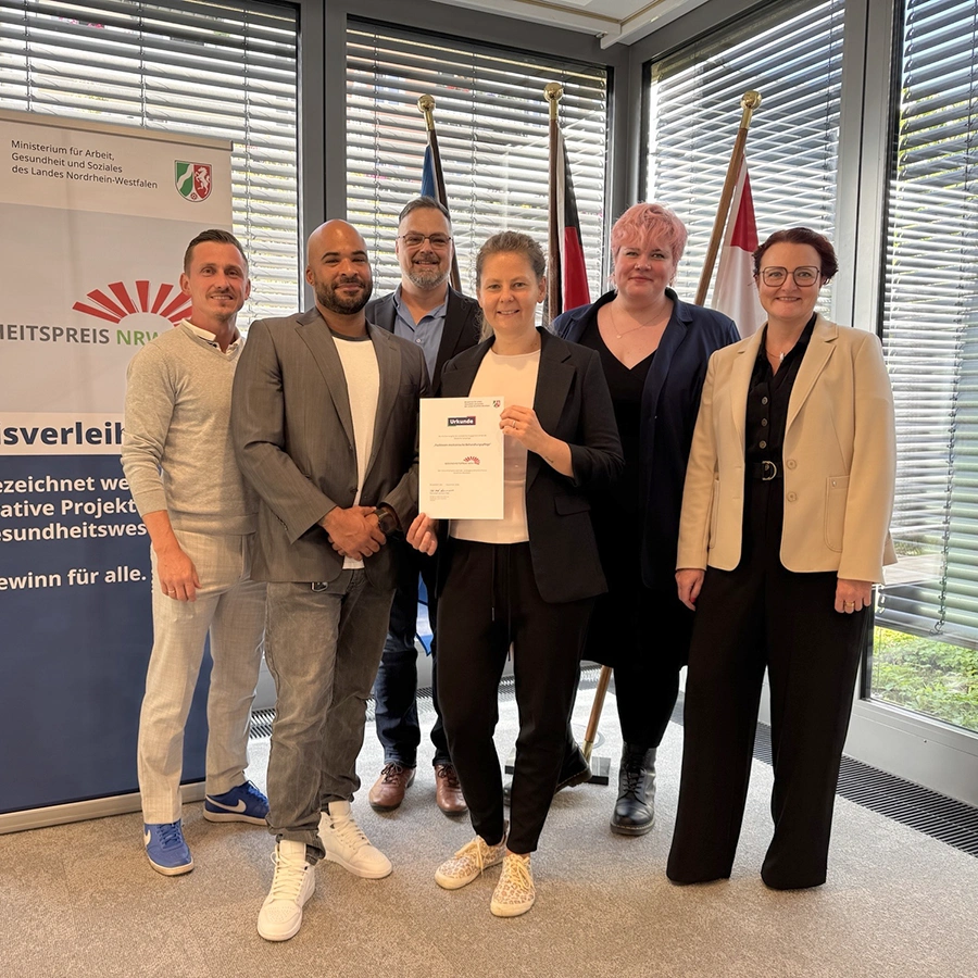 6 Personen stehen vor einem Plakat zur Preisverleihung des NRW Gesundheitspreises, wobei eine in der Mitte die dazugehörige Urkunde mit beiden Händen hochhält. Alles lächeln in die Kamera.
