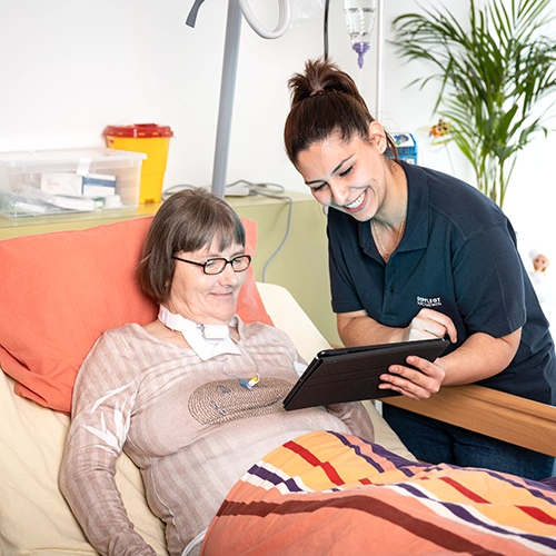 Weibliche Pflegekraft steht vornübergebeugt an einem Bett mit einer Klientin und zeigt ihr etwas auf einem Laptop. Beide lächeln.