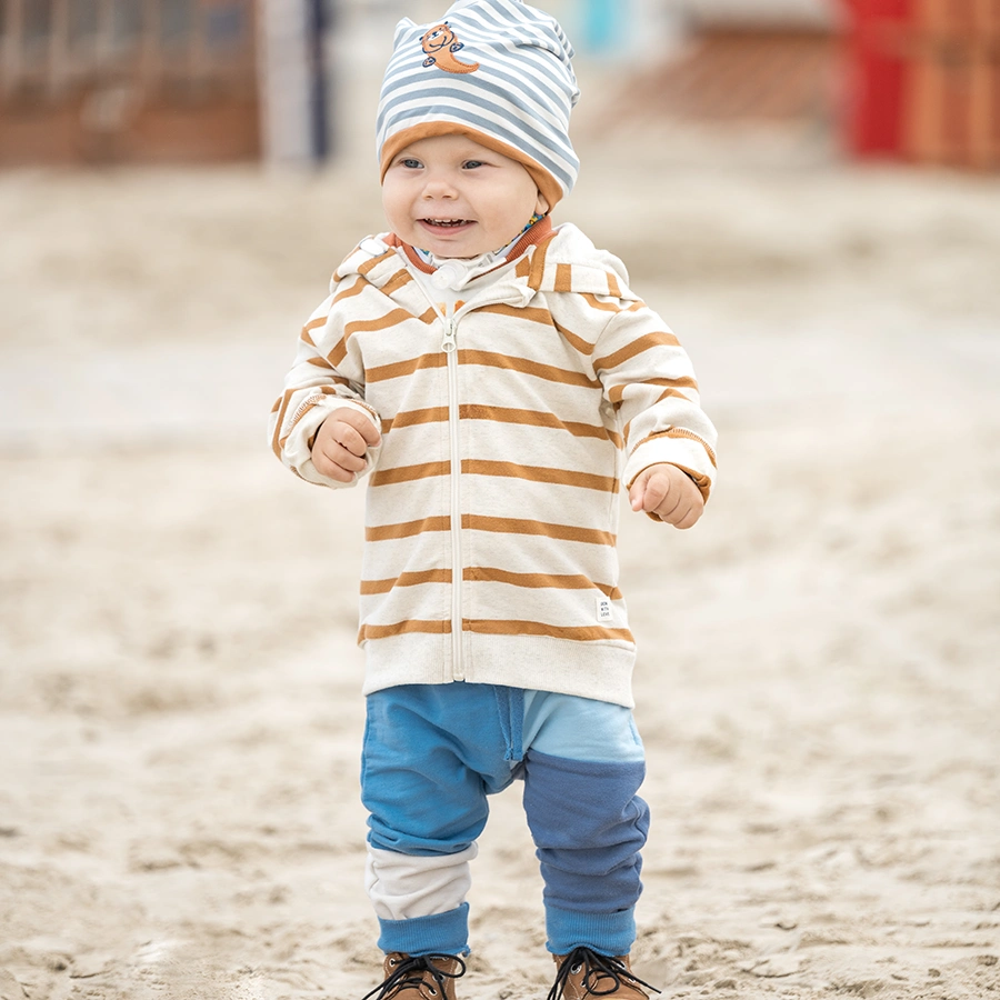 Kleiner Junge steht im Sandkasten, lächelt dabei an der Kamera vorbei und ist auf etwas anderes fokussiert.