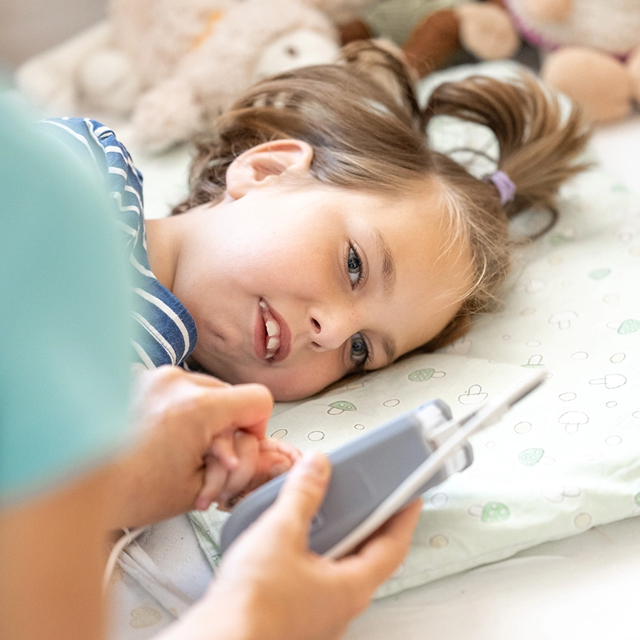 Kind liegt auf der Seite im Bett, im Fokus das Gesicht. Vor ihr angeschnitten und leicht verschwommen eine Pflegekraft mit einem kleinen Messgerät in der Hand.