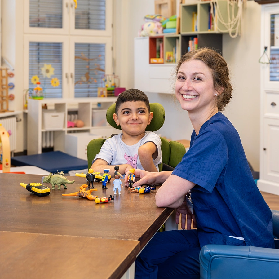 Kind im Rollstuhl lächelt eine weibliche Pflegekraft an. Die Pflegekraft lacht in die Kamera. Beide sitzen in einem Aufenthaltsraum für Kinder an einem Tisch, auf dem Spielfiguren stehen.