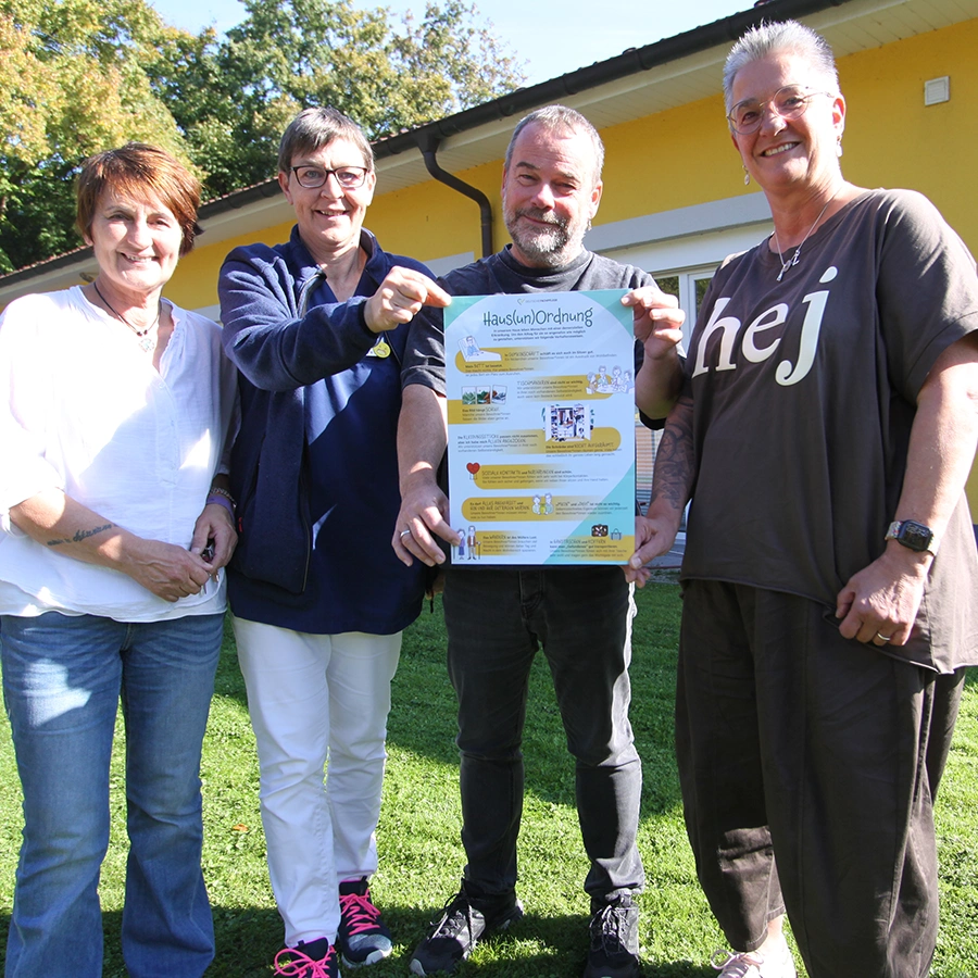 4 Mitarbeiter*innen stehen nebeneinander vor dem Gebäude auf dem Rasen. Alle lächeln in die Kamera. Beide Mitarbeiter*innen in der Mitte halten ein Plakat hoch.